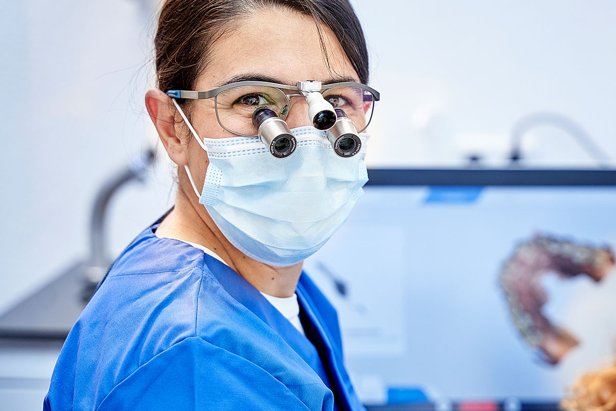 CMD-Behandlung in Hückelhoven: Zahnärztin lacht mit Lupenbrille und Maske in die Kamera.