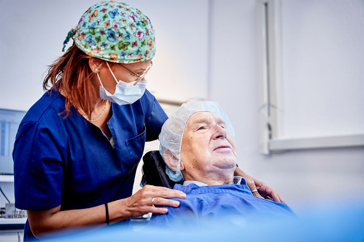 Älterer Herr im Zahnarztstuhl, eine ZFA legt die Hände auf seine Schulter und spricht ihm Mut zu für den Knochenaufbau in der Zahnarztpraxis Kowadent Hückelhoven